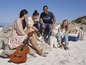 Een groep vrienden die op het strand chillen met een gitaar, een hond en een koelbox.