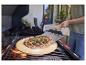 Pizza op een pizzasteen op een grill, met een persoon die een pizzaschep gebruikt.