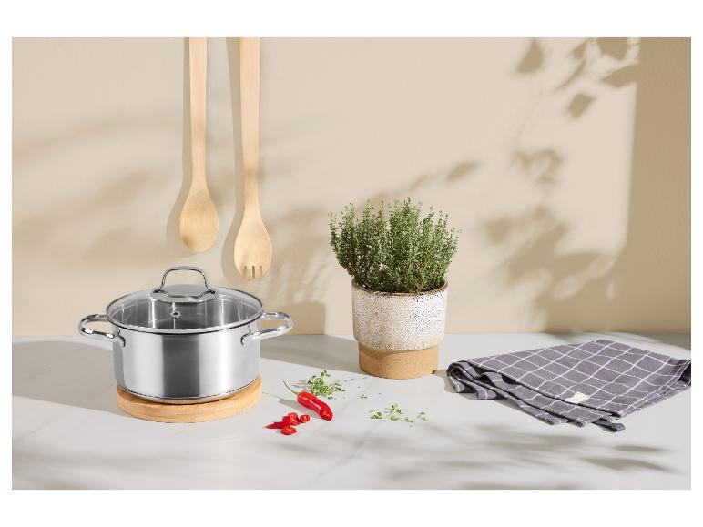 Roestvrijstalen pan met glazen deksel op houten onderzetter, met keukengerei en kruiden.