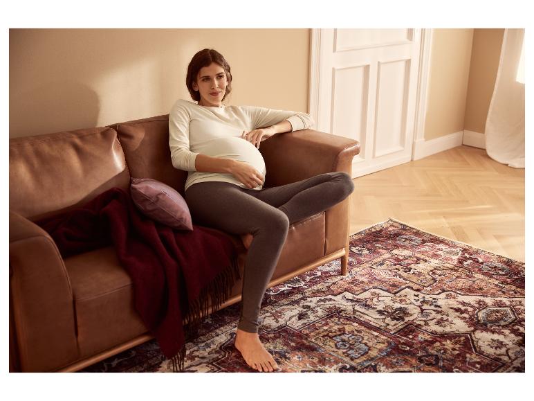 Een zwangere vrouw in een witte top met lange mouwen en grijze legging zit op een bruine leren bank.