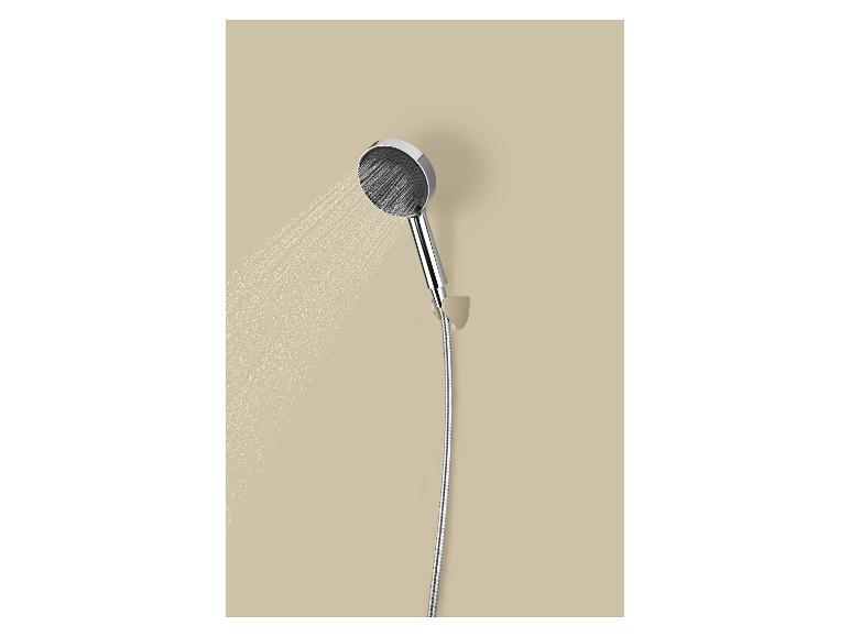 Chromen handdouche met slang die water sproeit tegen een beige muur.