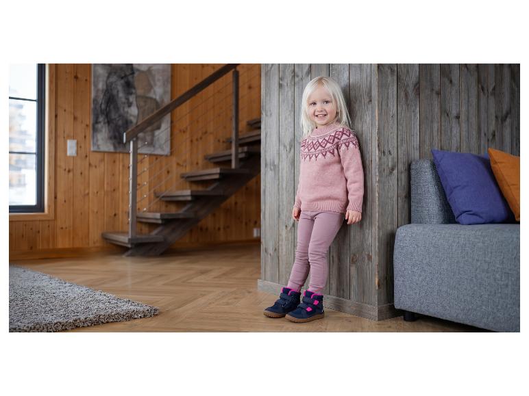 Lachend meisje in roze trui en broek, met schoenen aan, leunend tegen een houten muur.