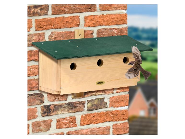 Houten vogelhuisje met groen dak gemonteerd op een bakstenen muur, met een vliegende vogel.