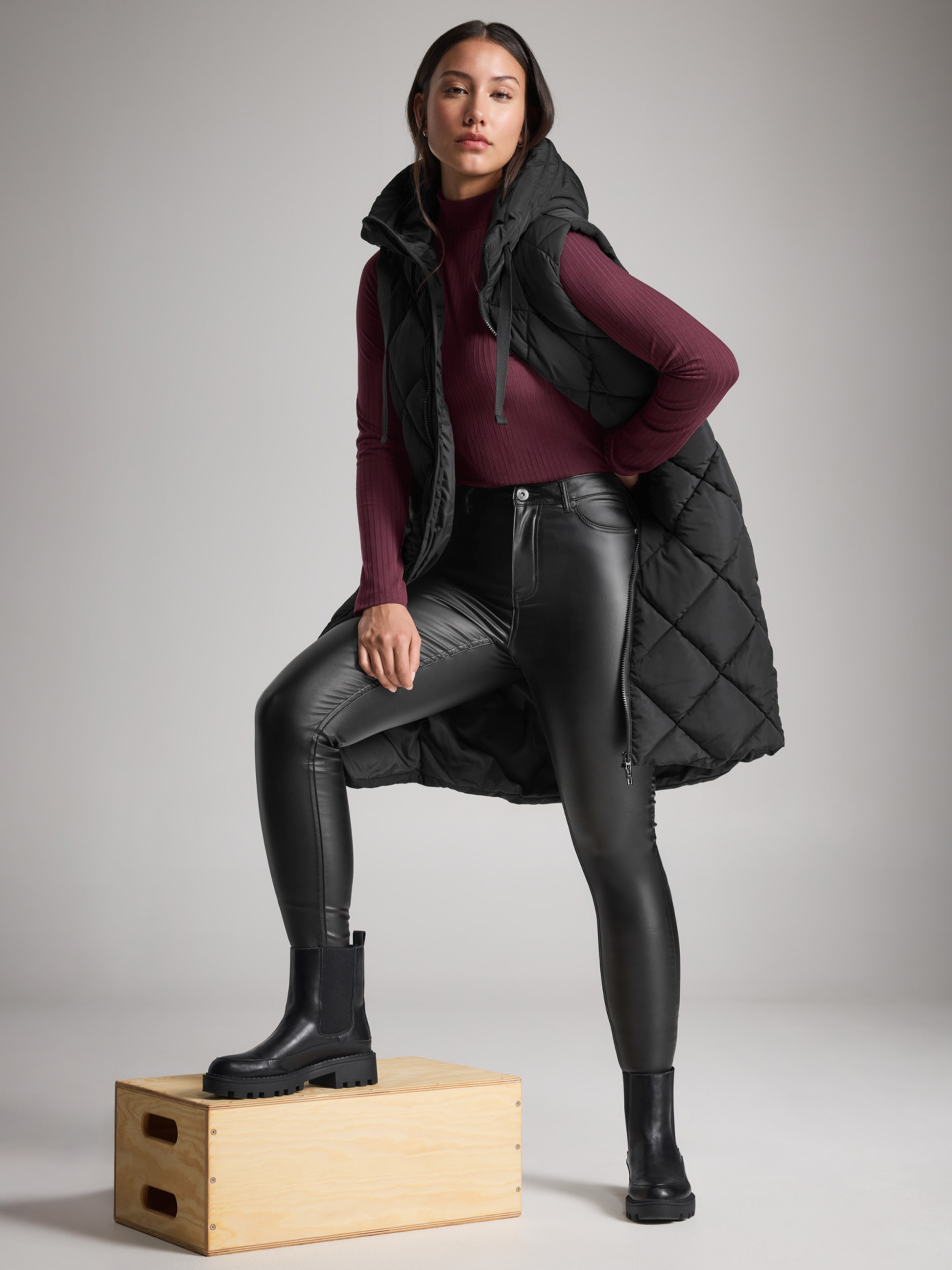 Vrouw in een zwart gewatteerd vest, leren jeans en laarzen, met één voet op een doos.