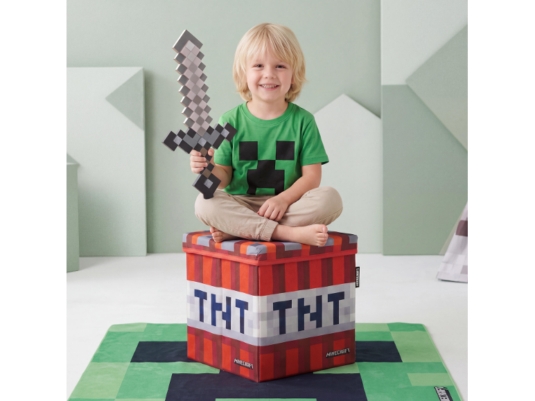Lachend kind met speelgoedzwaard, zittend op een TNT-blokvormige opbergdoos, op een videogame-thema vloerkleed.