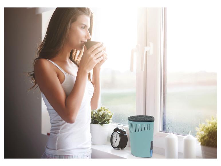 Een vrouw drinkt koffie bij een beslagen raam, met een luchtontvochtiger op de voorgrond.