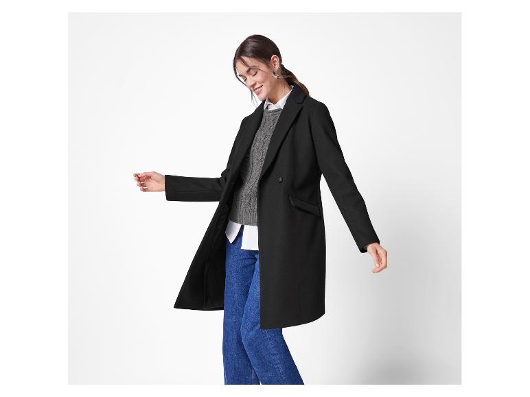 Een vrouw modelleert een zwarte, knielange jas over een grijze trui en een blauwe jeans.