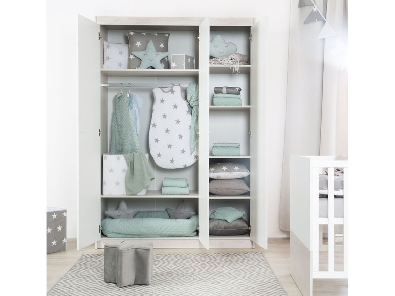 Een babykleedkamer met een witte kast vol met babykleding, een slaapzak en een babybedje.