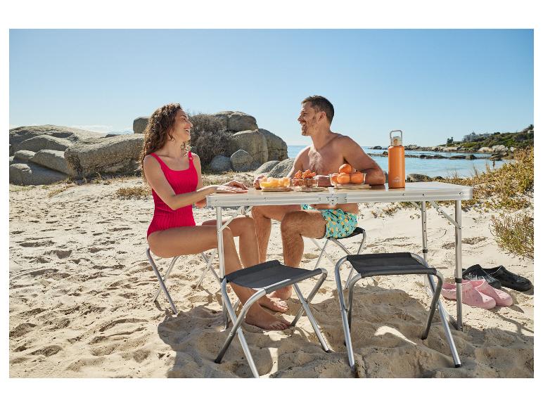 Koppel in zwemkleding picknickt op het strand met een opvouwbare tafel en stoelen.