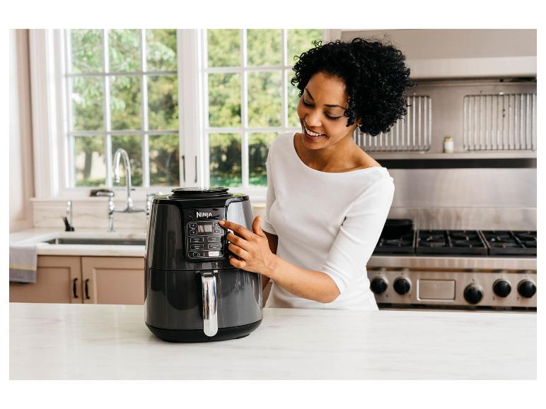 Vrouw in de keuken gebruikt een airfryer, lachend en kijkend naar het apparaat.