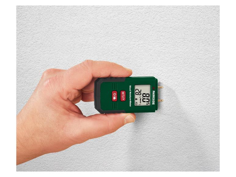 Hand houdt groene en zwarte houtvochtmeter vast die 28°C en 8.0% weergeeft op een witte muur.