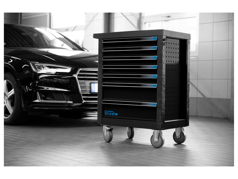 Een zwarte gereedschapswagen met meerdere laden, merk Güde, met een auto op de achtergrond.