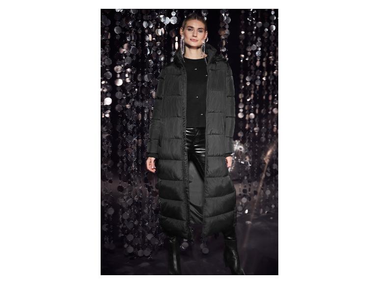 Vrouw in een lange zwarte jas, leren broek en laarzen, tegen een glimmende achtergrond.