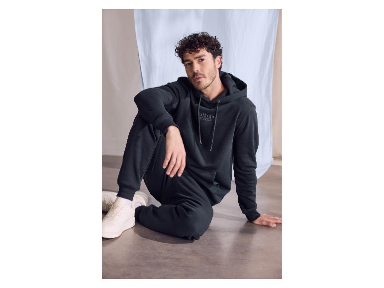 Man in een donkerblauwe hoodie en joggingbroek, zittend op de vloer.