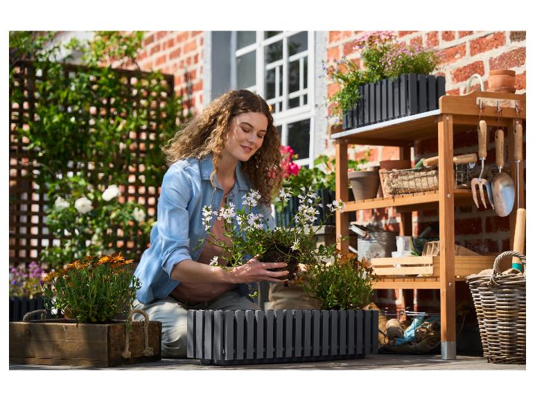 Vrouw plant bloemen in een grijze plantenbak op een houten terras met tuingereedschap.