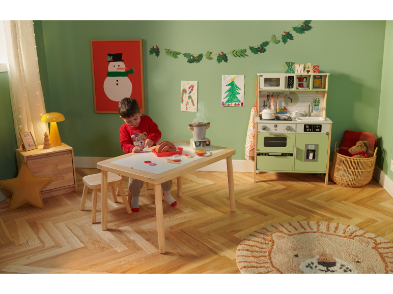 Kind speelt met een houten speelkeuken en speelgoedeten in een kerstversierde kamer.