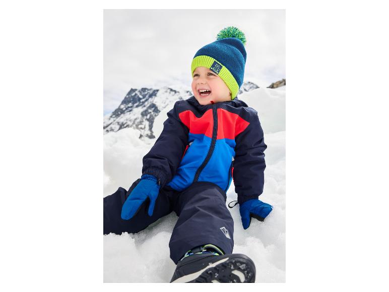 Blij kind in skipak en muts met pompon, zittend in de sneeuw.