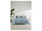 Slaapkamer met blauw beddengoed met patroon, houten hoofdbord, plant en bijzettafel.