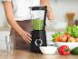 Een vrouw bereidt een groene smoothie in een blender, met vers fruit en groenten op tafel.