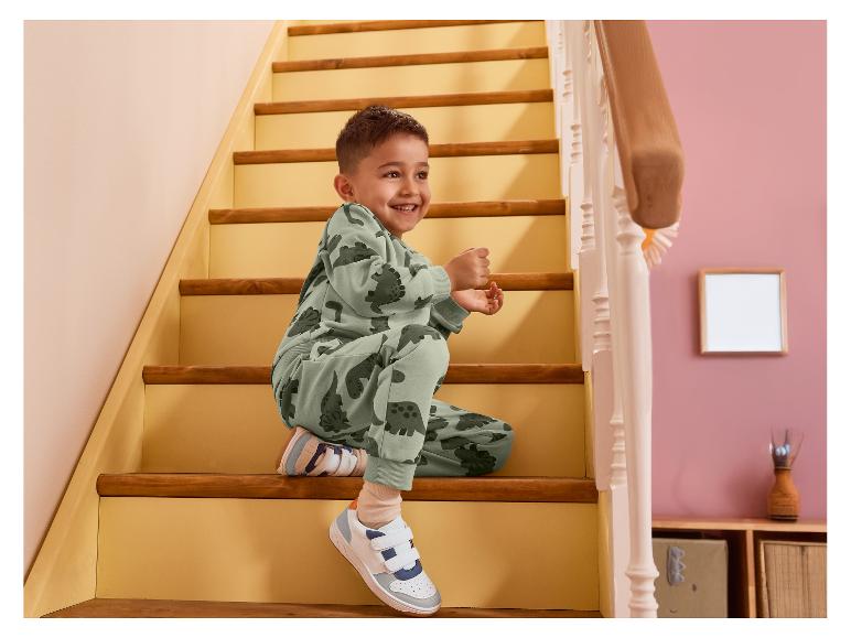 Lachende jongen in groen dinosaurus trainingspak zit op de trap.
