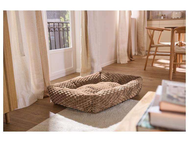 Beige dierenmand op een pluizig tapijt in een woonkamer met raam en stoelen.