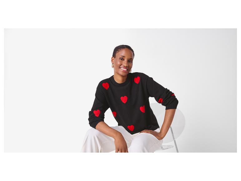 Vrouw in zwarte trui met hartjespatroon en witte broek, zittend op een stoel.