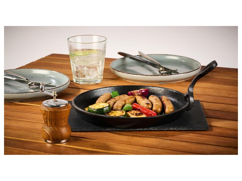 Gietijzeren pan met worstjes en gegrilde groenten op een houten tafel.