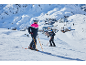 Twee skiërs in skikleding en helmen op een besneeuwde berghelling met een dorp op de achtergrond.