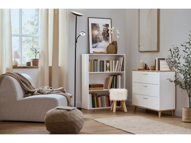 Gezellige woonkamer met een beige fauteuil, gebreide poef en witte boekenkast.