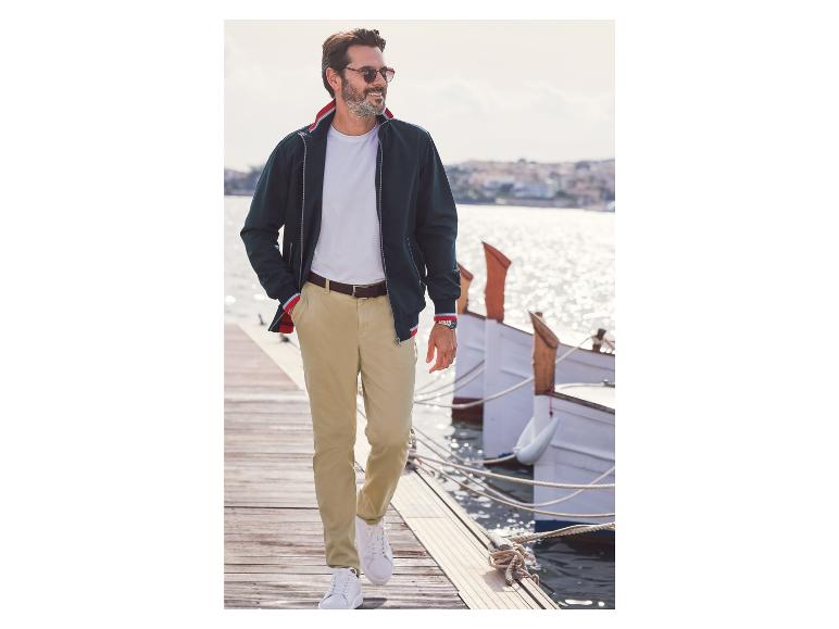 Man in een blauwe jas, beige broek en witte sneakers op een pier.