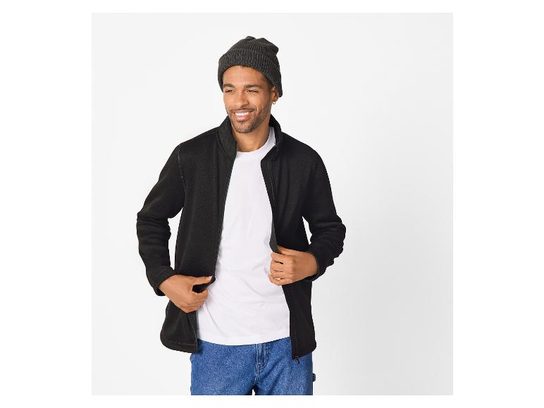 Man in een zwart fleecejack, wit T-shirt, blauwe jeans en een donkere muts.