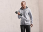 Man in een grijs hoodie met rits en zwarte broek, hij tilt een haltergewicht op.