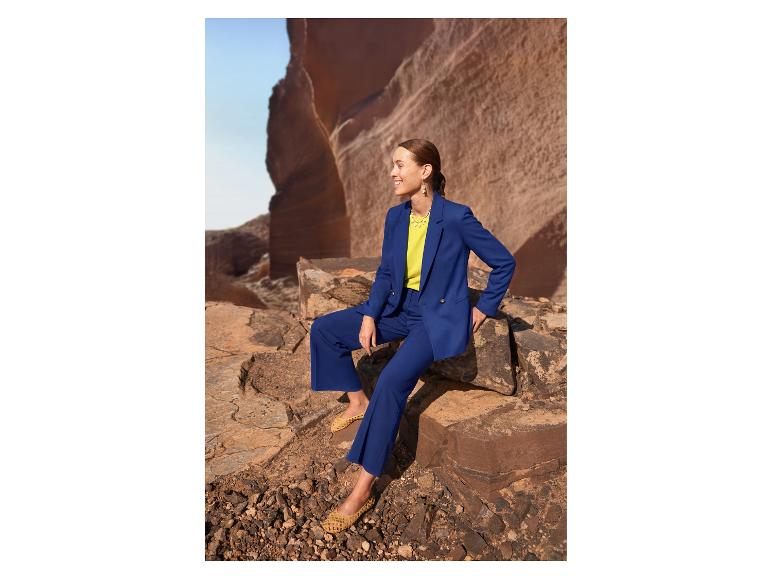 Vrouw in blauw pak en gele top, zittend op rotsen in een woestijnlandschap.