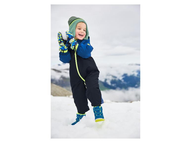 Blij kind in blauw skipak en groene muts speelt in de sneeuw.