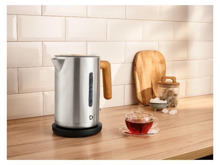 Roestvrijstalen waterkoker met houten handvat, een kopje thee en theeaccessoires op een houten aanrecht.