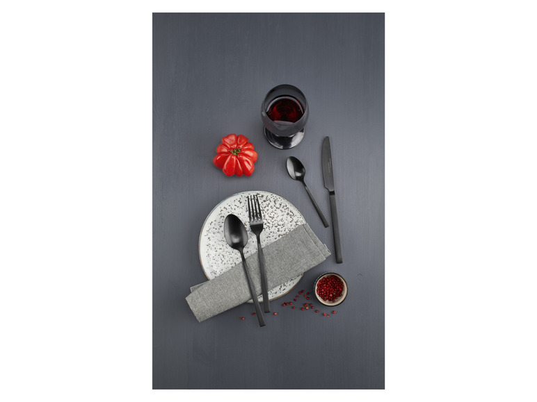 Zwart bestek, een bord, servet, een glas rode wijn en peperkorrels op een donkere houten tafel.