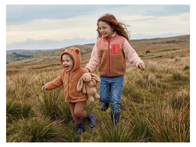 Twee blije kinderen in fleece jassen en regenlaarzen rennen door een veld.