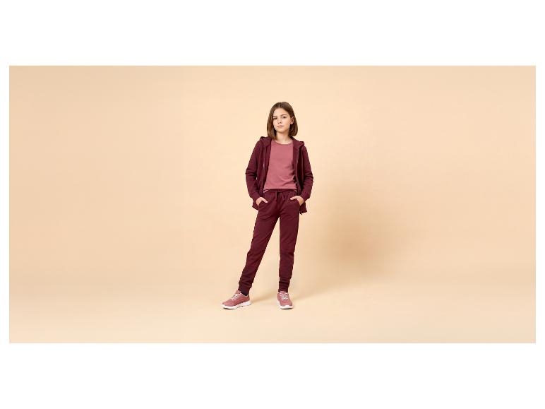Meisje in bordeauxrood trainingspak en roze T-shirt, poserend met handen in zakken.