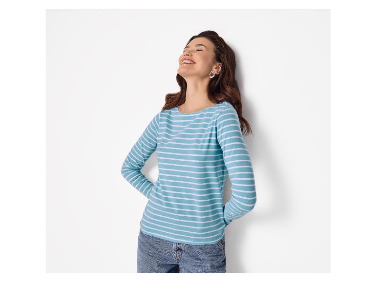 Vrouw in een blauw gestreepte top en jeans, lachend, leunend tegen een muur.