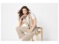 Vrouw in een beige gilet en broek, zittend op een houten kruk.