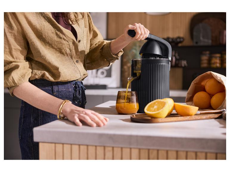 Vrouw perst sinaasappels met een zwarte elektrische citruspers, sap in een glas.