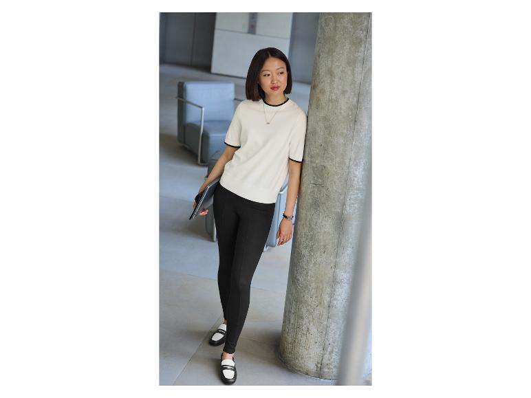 Vrouw in een crèmekleurige blouse en zwarte broek leunt tegen een betonnen pilaar.