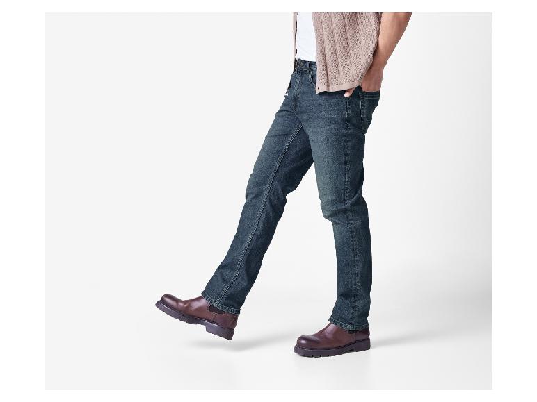 Een man in donkere jeans, een wit t-shirt en een lichtbruin vest.