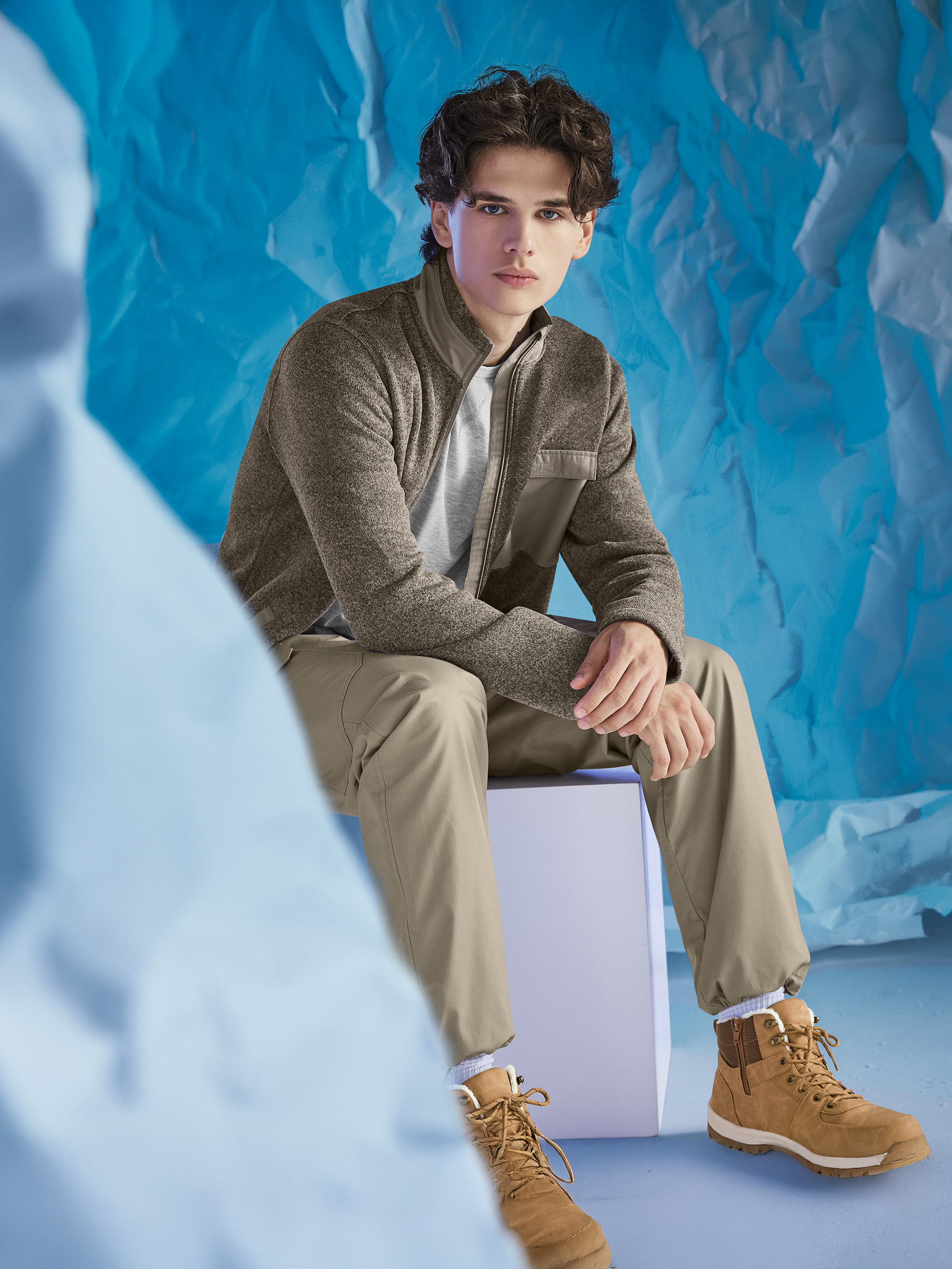 Man draagt een bruine jas, grijs T-shirt en kaki broek, zittend tegen een blauwe achtergrond.