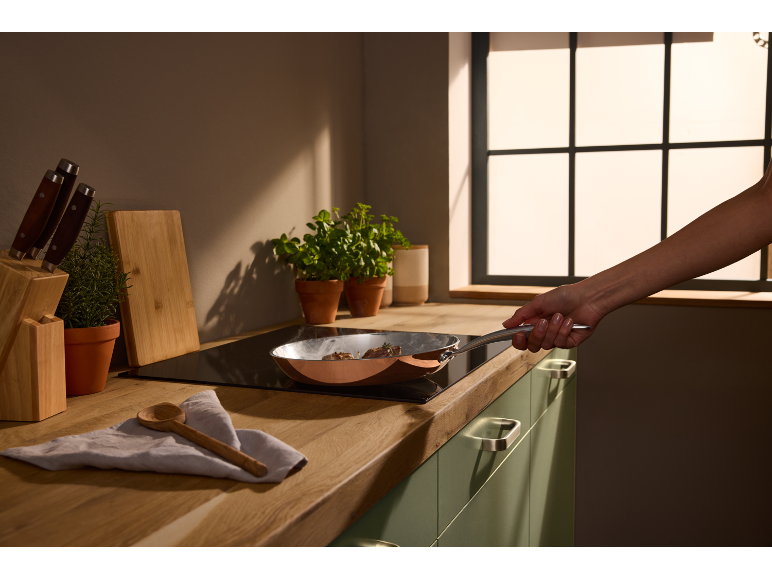 Persoon kookt vlees in een koperen pan op een inductiekookplaat in een moderne keuken.