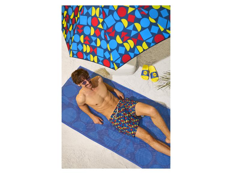 Man op strandmat in zwembroek en zonnebril onder parasol, met slippers ernaast.