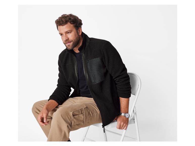 Man in zwart fleecejack en kaki broek zit op een witte stoel.