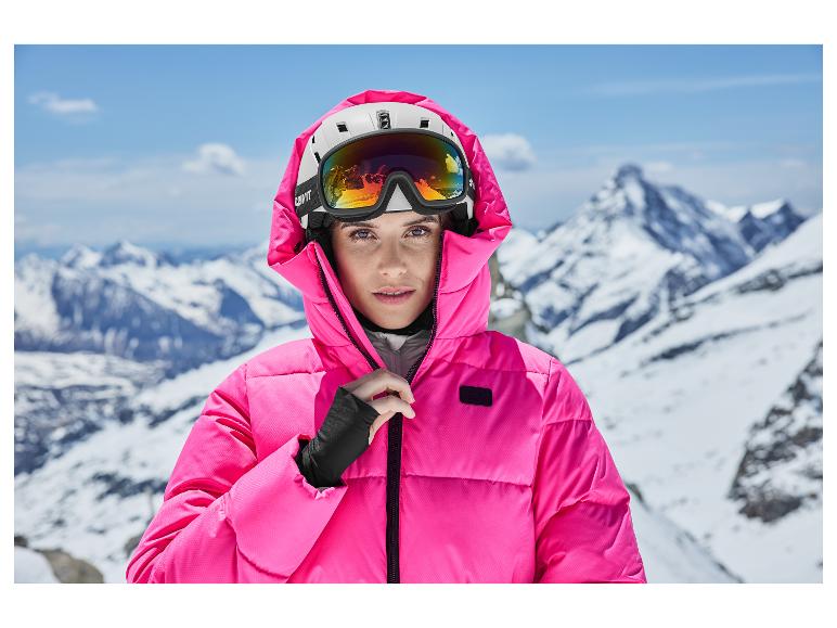 Vrouw in roze ski-jas en helm met skibril tegen besneeuwde bergachtergrond.