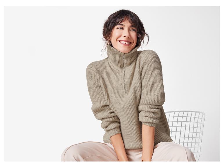 Vrouw zit op een stoel met een beige trui met rits en lichtroze broek.