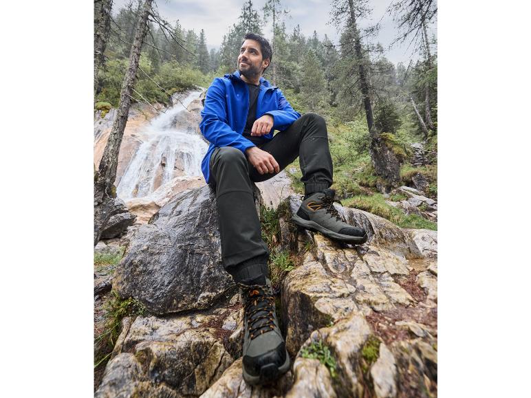 Man in een blauwe jas en zwarte broek in de buurt van een waterval, met zwarte wandelschoenen.